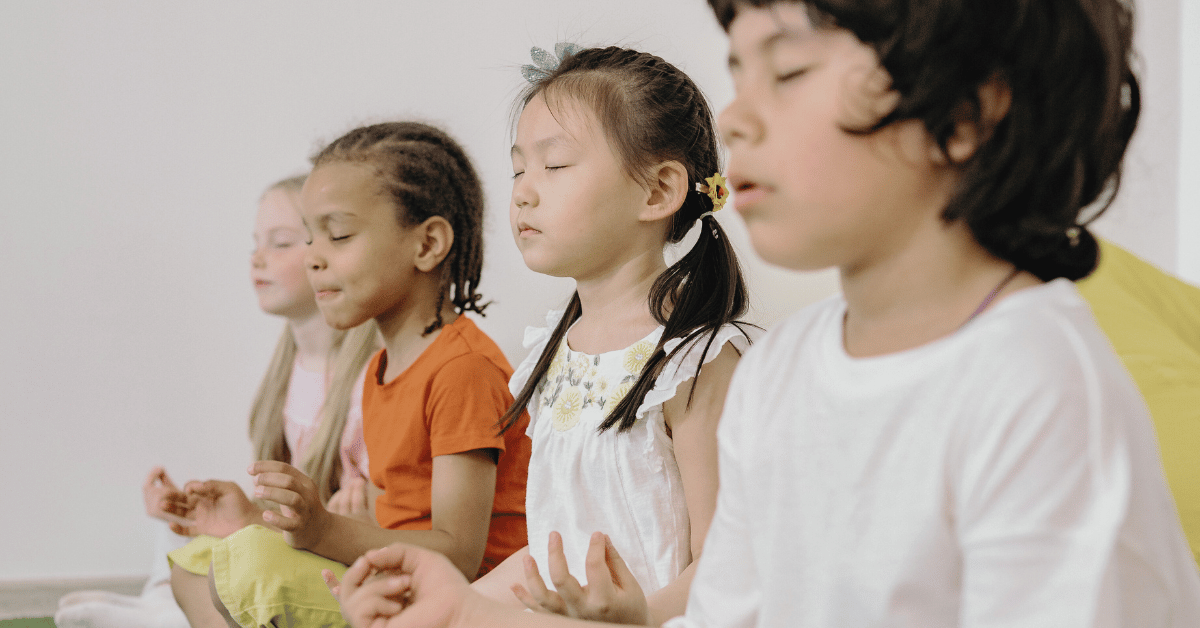**Alt text:** Four young children sit side-by-side on the floor with eyes closed and hands resting on their knees, appearing calm and focused during a mindfulness or meditation activity in a bright room.