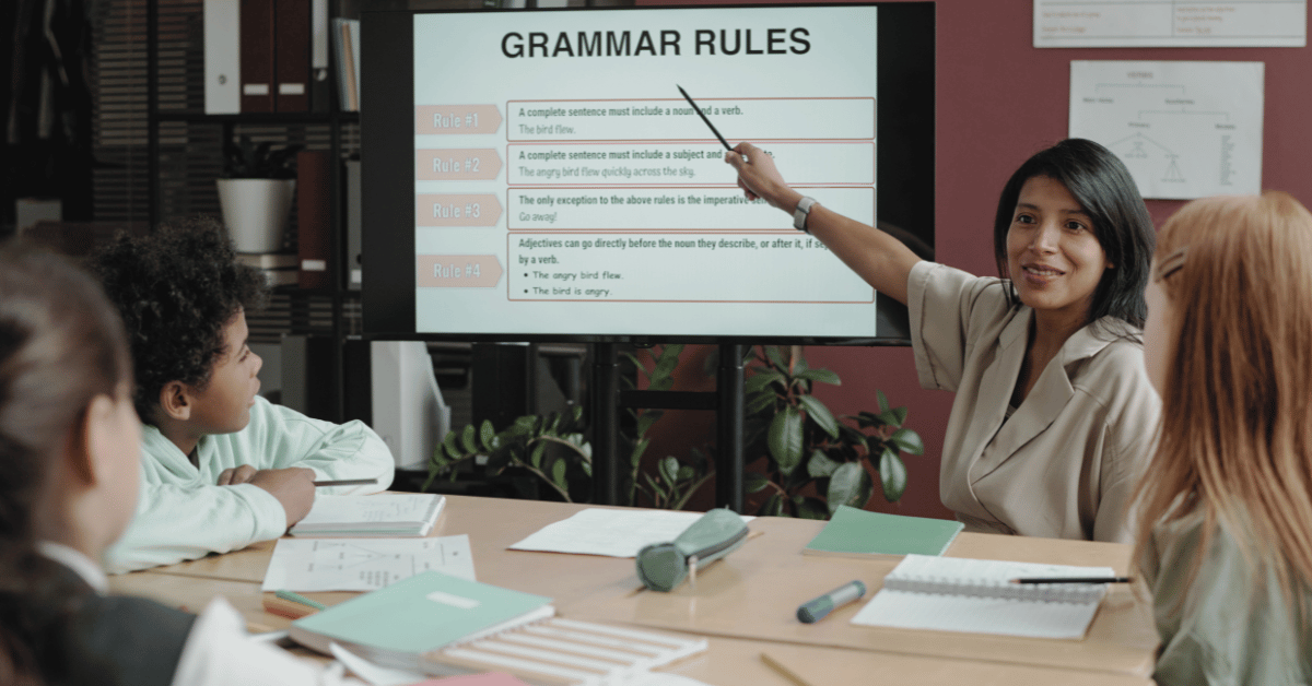 **Alt text:** A teacher points to a screen labeled “Grammar Rules” while sitting at a table with several young students, who are looking at the display and their notebooks during a small-group classroom lesson.